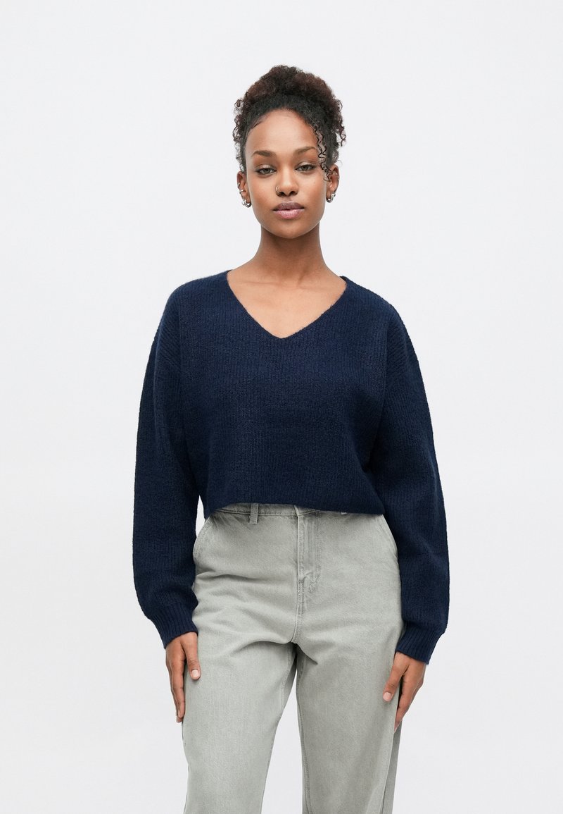 Pull en tricot bleu marine avec un col en V et des manches longues, associé à un pantalon taille haute gris clair. Les matériaux texturés sont visibles.