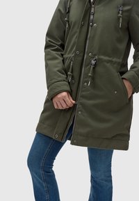 Parka verde oliva con cierre de cremallera y botones a presión, capucha con cordón, dos bolsillos laterales y puños acanalados. Lleva puesta sobre unos vaqueros azules.