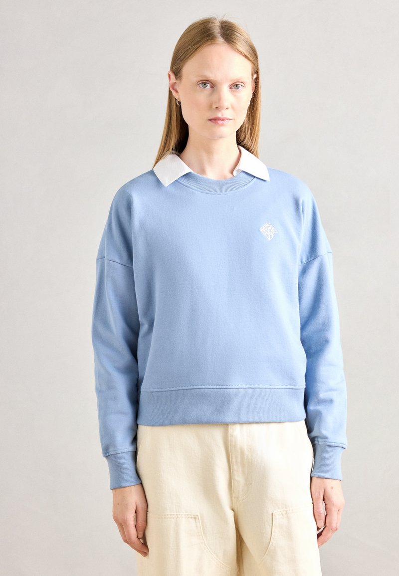 Sweatshirt bleu clair avec un col rond, des poignets et un ourlet côtelés. Présente une chemise blanche à col en dessous et un logo brodé.