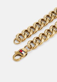 Goldene Kette mit dicken, miteinander verbundenen Gliedern und einer glatten, polierten Oberfläche. Enthält einen Karabinerhaken mit einem farbigen Emblemdetail.