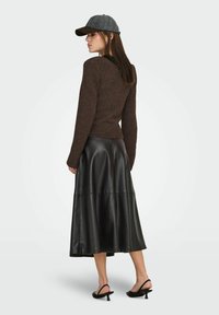 Pull en maille marron, jupe midi en cuir noir, et casquette de baseball grise. La jupe présente un design fluide avec des coutures verticales. Sandales à talons noires.