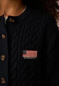 Jūras spēku adīts kardigans ar lielām brūnām pogām un uzšuvi ar Amerikas karogu un "SUPERDRY" logo. Tekstūras adījuma raksts.