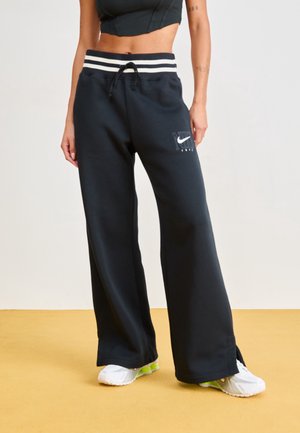 Kvinde iført sorte Nike sweatpants med brede ben, hvidt linjet taljebånd og hvide sneakers, stående på gul gulv.