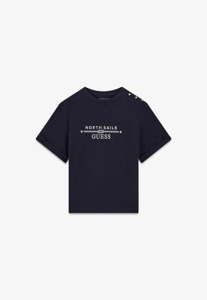 Camiseta azul marino de manga corta con puños doblados, logo blanco "North Sails Guess" centrado y tres botones dorados en el hombro izquierdo.