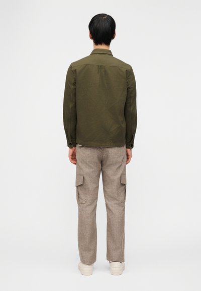 Άνδρας που φοράει χακί μπουφάν, μπεζ παντελόνι cargo και λευκά αθλητικά παπούτσια, στέκεται με την πλάτη στραμμένη σε απλό λευκό φόντο.