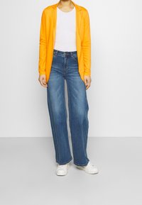 Cardigan lungo arancione con maniche lunghe sopra una camicia bianca, abbinato a jeans a gamba larga blu e sneakers bianche. Outfit casual con textures sovrapposte.