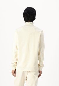 Svijetla krem majica sa visokim ovratnikom, rebrastim manžetama i porubom. Glatka tekstura s opuštenim krojem; minimalno brendiranje na rukavu.