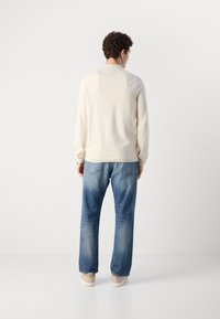 Beige long-sleeve shirt with ribbed texture, paired with faded blue jeans and light-colored sneakers, seen from the back.