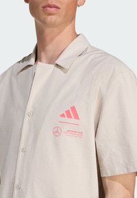 Jasno beżowa koszula z krótkim rękawem z teksturowanej tkaniny. Posiada różowe logo Adidas i Mercedes po lewej stronie. Zapięcie na guziki z przodu.