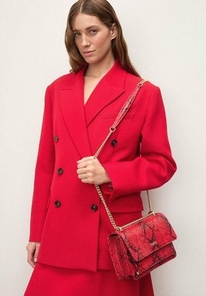 Femme portant un blazer croisé rouge et une jupe, tenant un sac à main à motif peau de serpent rouge avec une bandoulière chaîne dorée sur un fond clair.