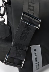 Borsa in nylon nera con una tracolla testurizzata dotata di una fibbia in metallo, dettagli in pelle e un'etichetta grigia con la scritta "KARL LAGERFELD JEANS."