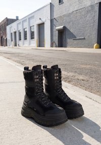 Pair of black lace-up platform boots with fleece lining placed on a concrete sidewalk in an urban street setting.