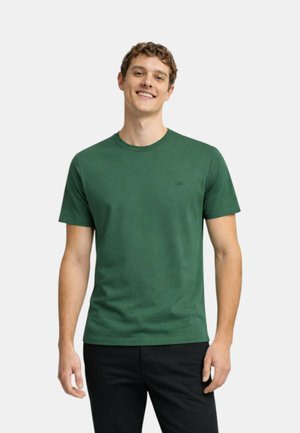 Giovane uomo sorridente, indossa una semplice maglietta verde scuro a maniche corte e pantaloni neri, in piedi davanti a uno sfondo bianco.