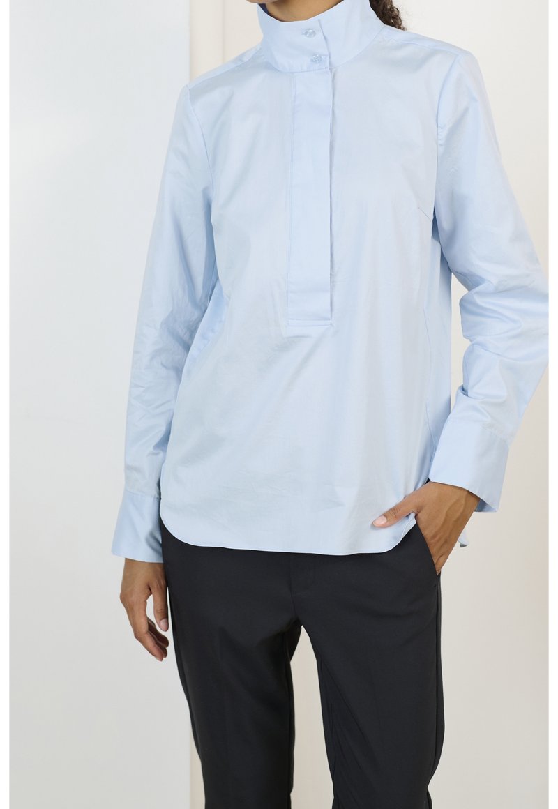 Femme portant une chemise bleu clair à manches longues avec un col montant et un pantalon noir, une main dans la poche, debout à l'intérieur.