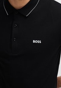 Svart polo t-shirt med en texturerad tyg, tre knappar och en vit broderad "BOSS"-logotyp på bröstet. Har en kontrasterande krage.