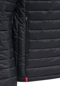 Détail d'une veste matelassée noire avec une poche zippée verticale et une petite étiquette de marque en tissu rouge en bas.