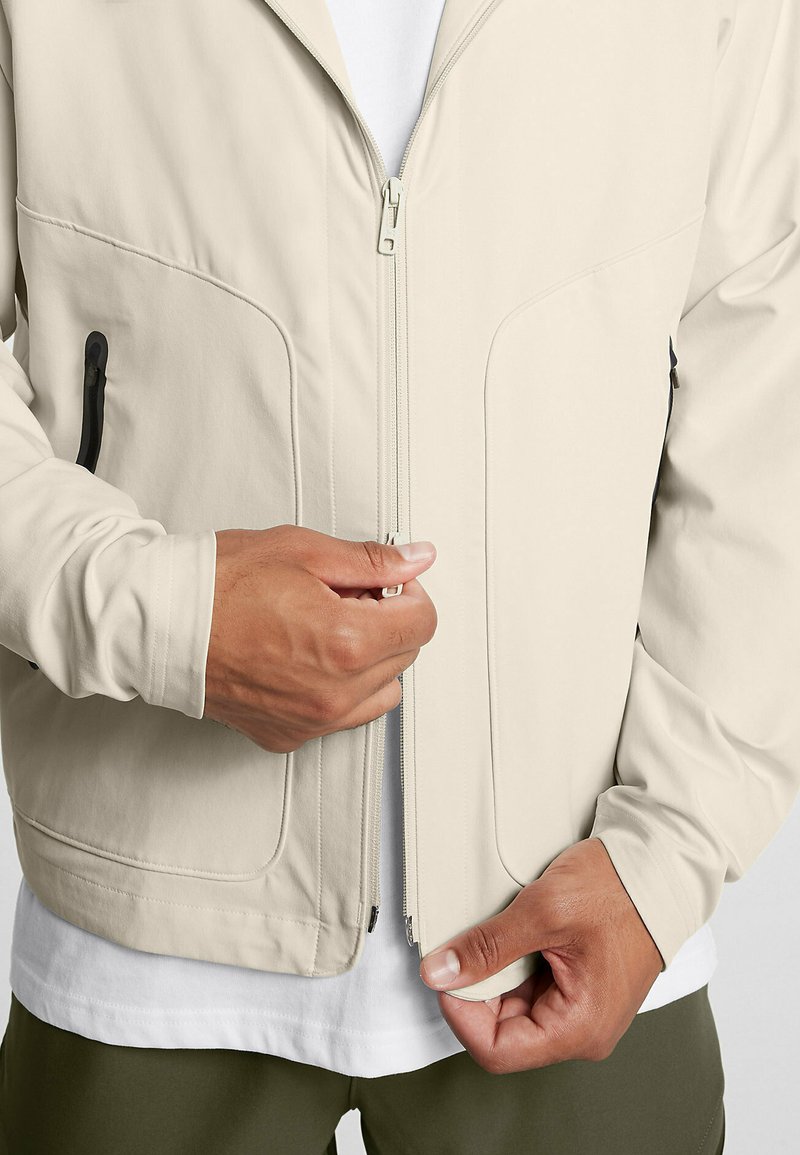 Veste zippée beige clair en tissu lisse, dotée de deux poches latérales, d'un col montant et de manches ajustées sur une chemise blanche.