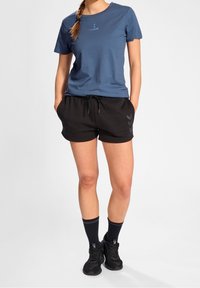 Modré bavlněné tričko s malým logem, spojené s černými elastickými šortkami s bočními pruhy a černými teniskami s žebrovanými ponožkami.