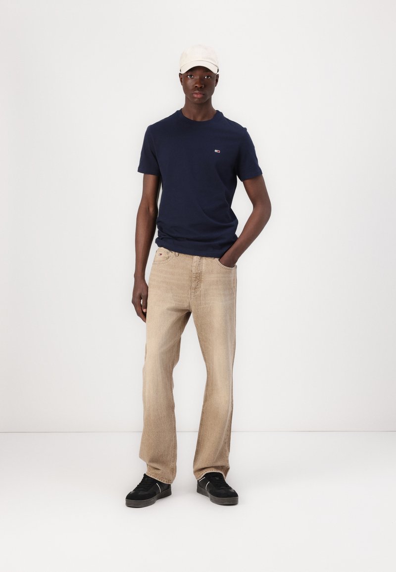 Tânăr purtând un tricou albastru închis, blugi bej, adidași negri și o șapcă albă, stând cu o mână în buzunar.