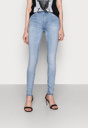Personne portant un jean skinny bleu clair, des talons hauts noirs ouverts aux orteils et un t-shirt noir avec un graphisme de carlin, debout devant un fond uni.