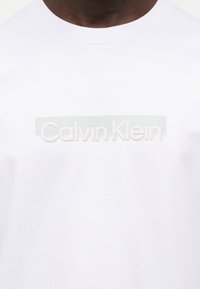 Κοντινό πλάνο ενός ατόμου που φοράει λευκό T-shirt με ανεπαίσθητο, ανάγλυφο λογότυπο "Calvin Klein" στο στήθος.