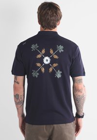 Homme tatoué portant un polo bleu marine avec un design floral, avec une marguerite blanche au centre et des feuilles vertes et orange croisées au dos.
