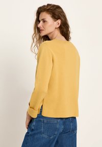 Frau mit welligem rotem Haar, die ein gelbes Langarmoberteil und blaue Jeans trägt, seitlich vor einem schlichten Hintergrund stehend.