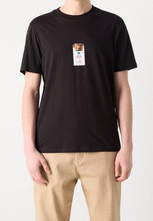 Mężczyzna w czarnej koszulce z małym nadrukiem głowy wielbłąda wystającej z białego opakowania oznaczonego napisem "Hugo" na piersi.