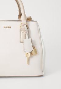 Sac à main structuré en matériau synthétique beige clair avec quincaillerie dorée. Équipé d'un accent clé et cadenas, finition lisse et poignées courtes.