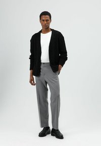 Zwarte kabelgebreide cardigan over een wit T-shirt, gecombineerd met grijze, op maat gemaakte pantalons en zwarte schoenen. Eenvoudig, gestructureerd ontwerp en pasvorm.