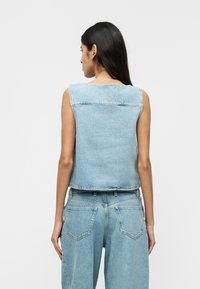 Femme aux longs cheveux foncés portant un gilet en jean bleu clair sans manches et un jean taille haute en jean bleu clair, vue de dos sur un fond uni.