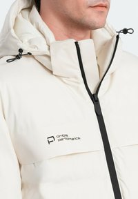 Vit isolerad jacka med hög krage, svart dragkedja och dragsnören. Har logotyp på bröstet och mjukt innerfoder.
