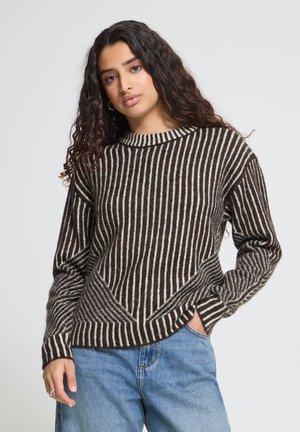 Femme aux longs cheveux bouclés portant un pull rayé marron et blanc et un jean bleu, debout avec une main dans la poche devant un fond uni.