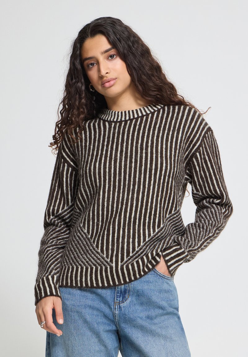 Femme aux longs cheveux bouclés portant un pull rayé marron et blanc et un jean bleu, debout avec une main dans la poche devant un fond uni.