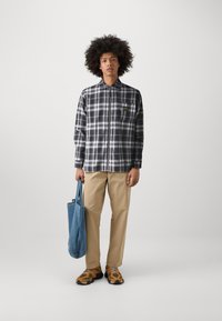 Checked black and white flannel shirt with a collar, paired with beige pants and brown/black sneakers. Blue tote bag in hand.