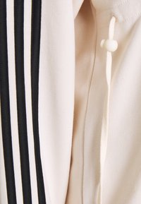Tessuto beige con tre strisce nere verticali e un cordino bianco con un fermacorda in plastica bianca su un indumento.