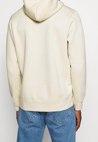 En person klädd i en krämfärgad hoodie och blå jeans, sedd bakifrån mot en enkel ljus bakgrund.