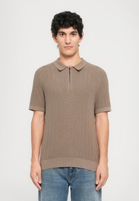 Ung mann med krøllete hår som har på seg en taupe kortermet ribbet polo genser med glidelås foran og blå jeans, stående mot en ensfarget bakgrunn.