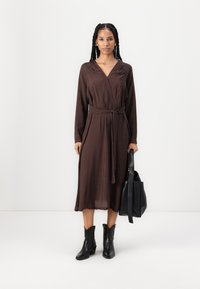 Robe enveloppante marron à manches longues avec ceinture à nouer, fabriquée en tissu léger, associée à des bottines noires et à un sac fourre-tout noir.