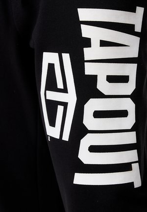 Schwarzer Stoff mit einem auffälligen weißen "TAPOUT"-Logo und geometrischen Akzenten, die an der Seite bedruckt sind. Glatte Textur und athletic Fit.