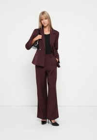 Burgundy színű, testre szabott blazer és azzal passzoló, bő szárú nadrág, fekete top és magassarkú cipő kombinációjában, amelynek szerkezete letisztult formákat és elegáns vonalakat mutat.