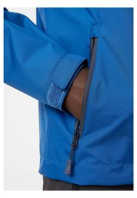 Blauwe jas met een klittenbandsluiting bij de mouw en een ritszak. De stof lijkt glad met een waterafstotende afwerking, evenals grijze accenten.