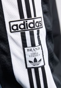 Černobílá sekce Adidas sportovní bundy s texturou síťoviny, obsahující tři svislé pruhy, nášivku s logem a značkovou etiketu.