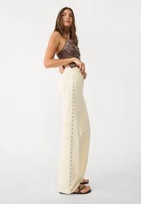 Cream wide-leg trousers with side button detail and back pockets, paired with a brown patterned cropped top. Flat sandals complete the look.