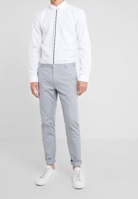 Valkoinen pitkähihaisen paita mustalla napituksella, yhdistetty vaaleanharmaisiin slim-fit-housuihin ja valkoisiin tennareihin. Huomaamaton tekstuuri näkyy.