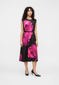 Robe noire avec de grandes imprimés floraux roses, manches courtes, taille cintrée avec une ceinture, et une jupe mi-longue fluide. Talons noirs pointus.