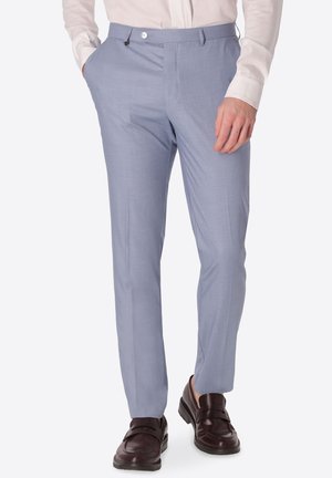 Uomo che indossa pantaloni azzurri su misura con camicia bianca, una mano in tasca e mocassini in pelle marrone scuro.