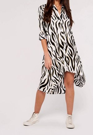Mujer con vestido de manga larga con estampado de cebra en blanco y negro con bajo asimétrico y zapatillas blancas de caña alta, de pie frente a un fondo liso.