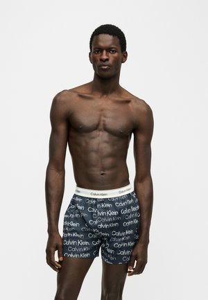 Fit man mit dunkler Haut, der marineblaue Boxershorts mit Calvin Klein-Logo trägt, steht vor einem schlichten hellen Hintergrund.