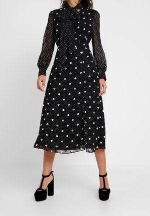 Black polka dot dress with sheer sleeves, tie-neck detail, mid-length hem, and black platform heels. Smooth, lightweight fabric.